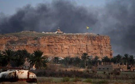 Smoke rises over the village of Baghouz behind flags of the People's Protection Units (YPG) and Women's Protection Units (YPJ) on March 22, 2019. Photo: Giuseppe Cacace/AFP