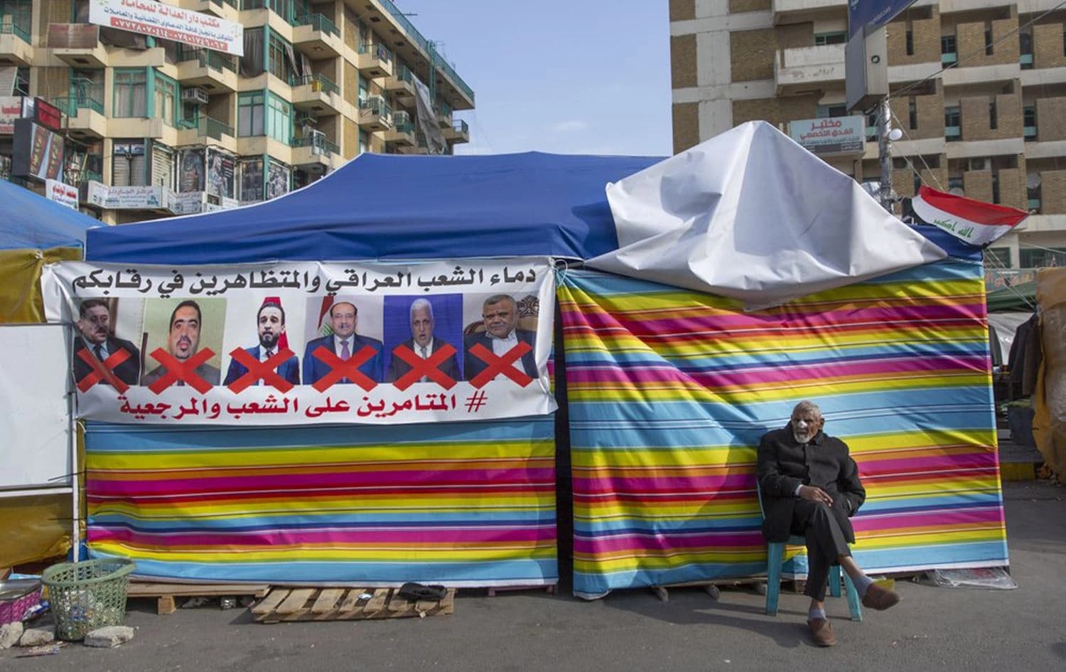 A protester sits next to a poster with defaced pictures of Iraqi politicians that reads, "the blood of the Iraqi people and the protesters is on your necks." Tahrir square,  Baghdad, Iraq, Wednesday, Dec. 25, 2019. Photo: Nasser Nasser/ AP 