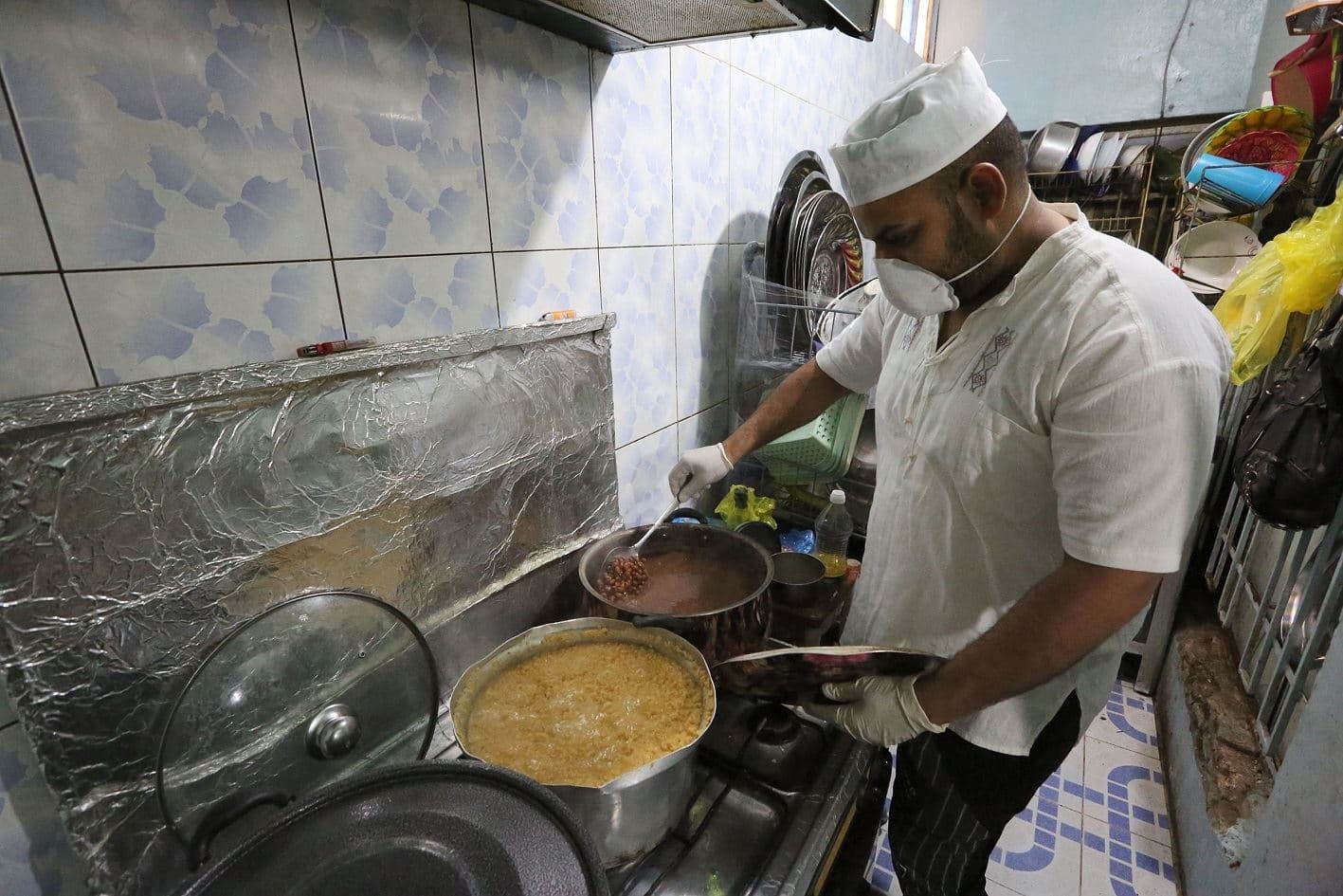 A mask-clad man cooks beans and other food for a family currently under lockdown due to COVID-19 coronavirus pandemic quarantine measures, in the Iraqi capital Baghdad on March 28, 2020. Photo: Sabah Arar 	