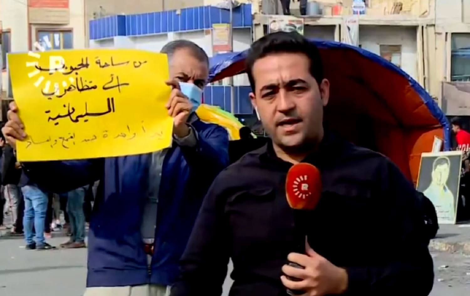 A protestor holds up a poster that reads: “From Habboubi square to Sulaimani protestors: one hand to fight violence and corruption” behind Rudaw’s Halkawt Aziz in Nasiriyah’s Habboubi Square on December 4, 2020. Photo: Rudaw