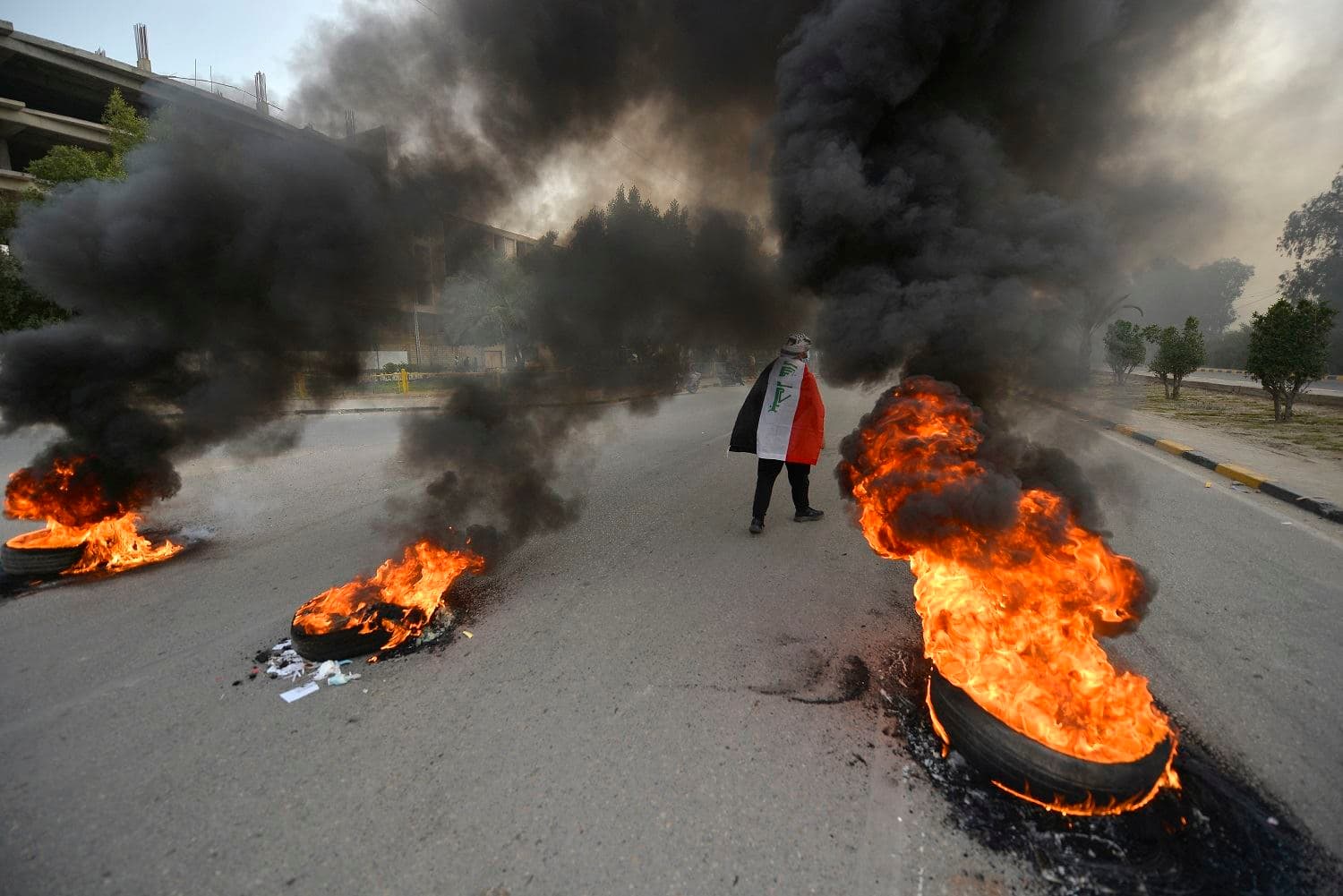 A demonstrator draped in the Iraqi national flag walks past flaming tires during clashes between anti-government protesters in Iraq's central holy shrine city of Najaf on March 15, 2021. Photo: Ali Najafi / AFP