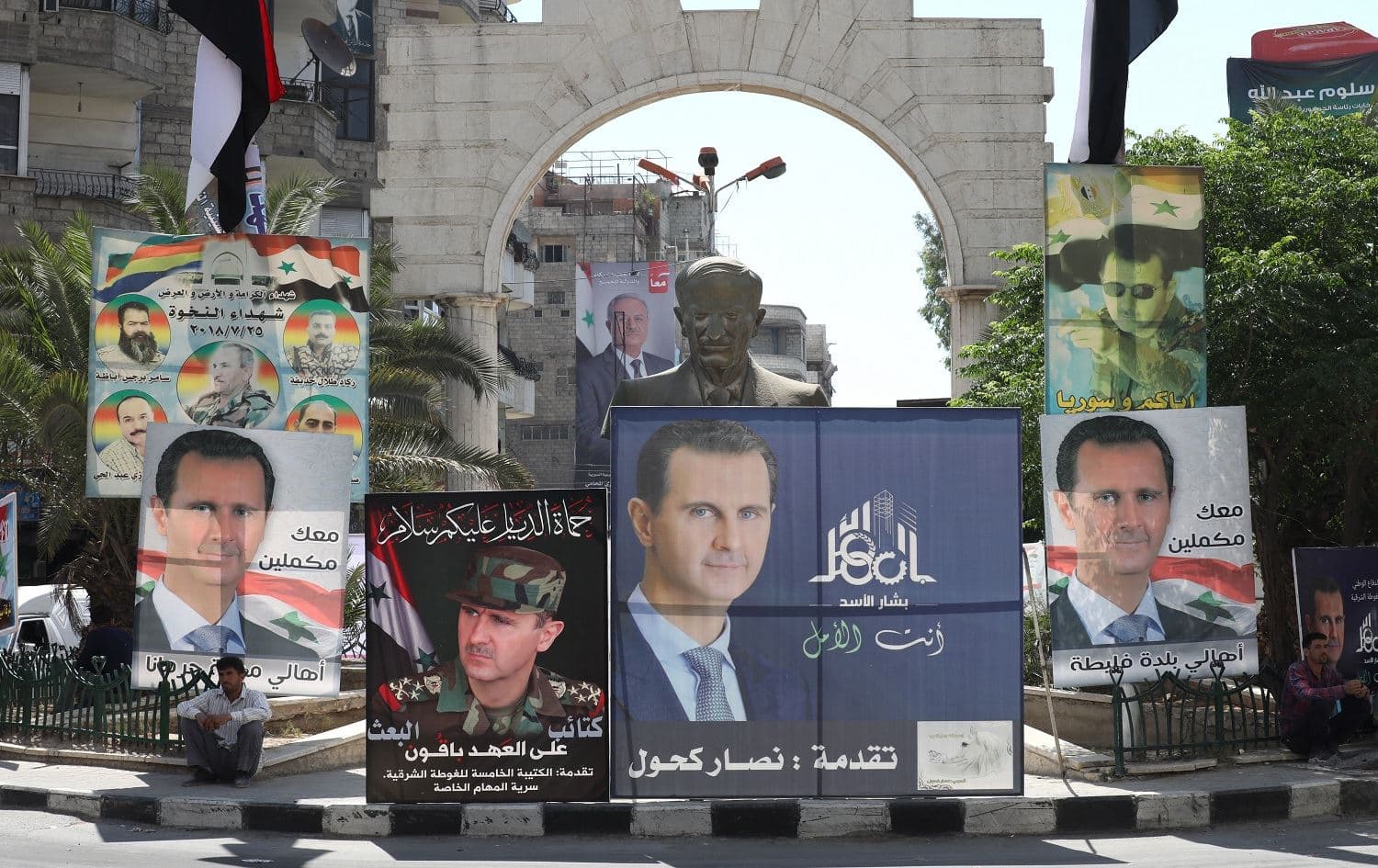 A man sits next to a statue of of Syria's late president Hafez al-Assad surrounded by campaign billboards depicting his son President Bashar al-Assad on May 23, 2021. Photo: Louai Beshara/AFP