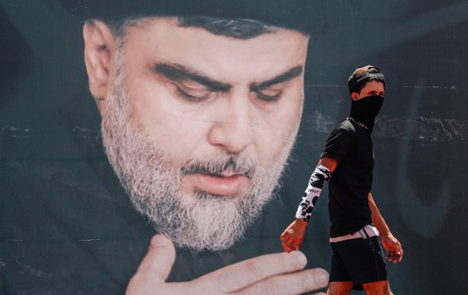 A mask-clad youth walks in front of a large poster of Iraq's populist Shiite Muslim cleric Muqtada al-Sadr in Sadr City, Baghdad on July 15, 2021. File Photo: Ahmad al-Rubaye/AFP