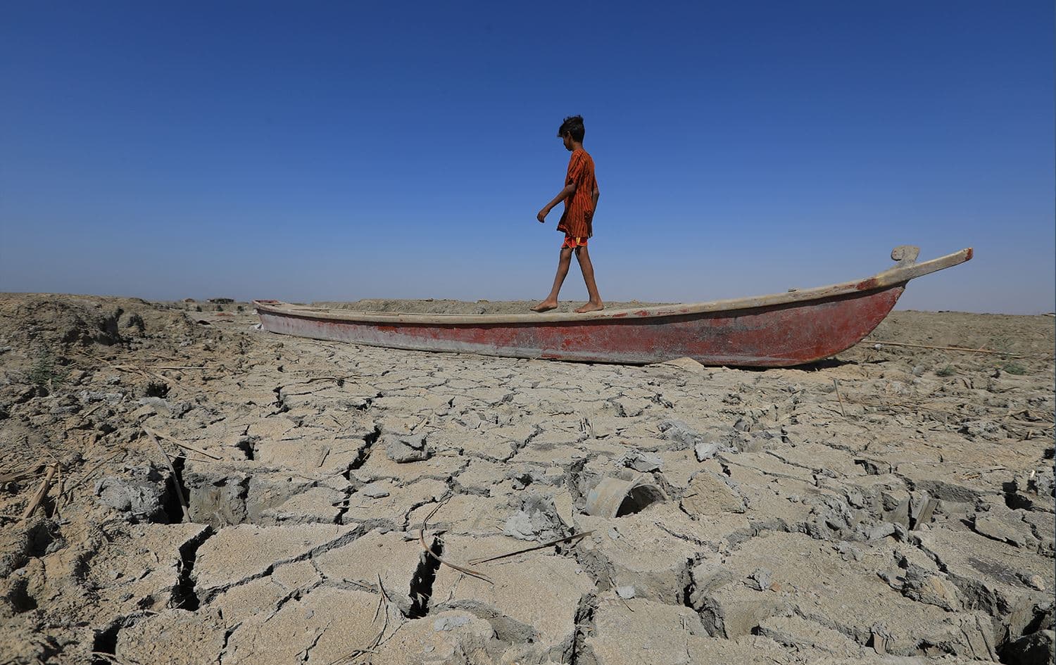 A boy walks on a boat left lying on the dried-up bed of a section of Iraq's receding southern marshes of Chibayish in Dhi Qar province, on June 28, 2022. Photo: Asaad Niazi/AFP 