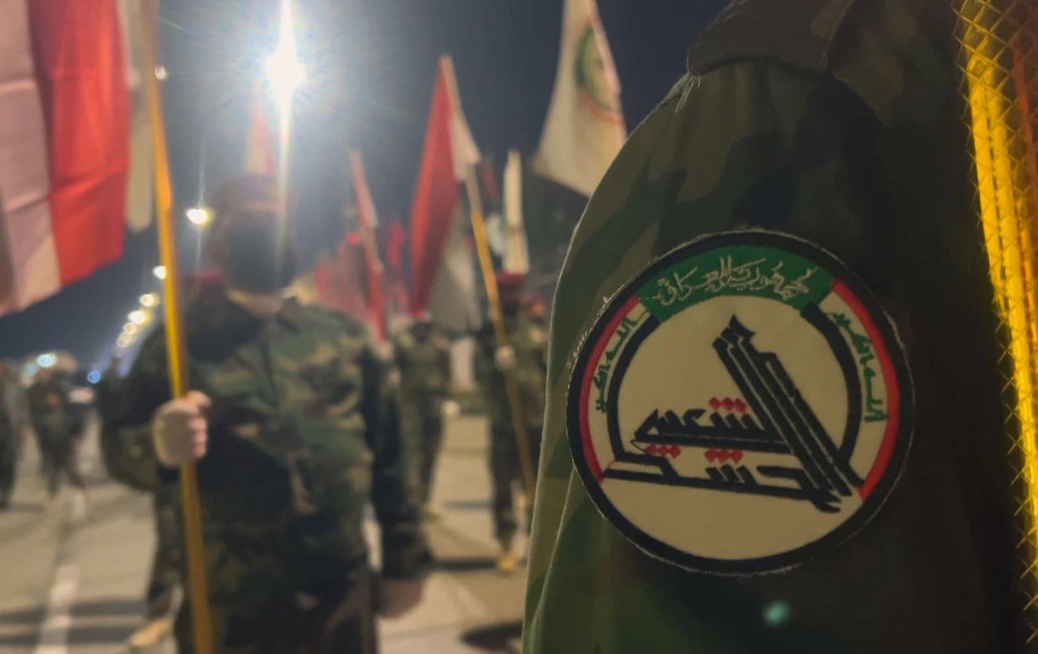 The PMF emblem on the arm of an Iraqi Shiite militiamen as others around him hold the Iraqi flag in a ceremony commemorating Soleimani and Muhandis on January 4, 2023. Photo: Rudaw/screengrab