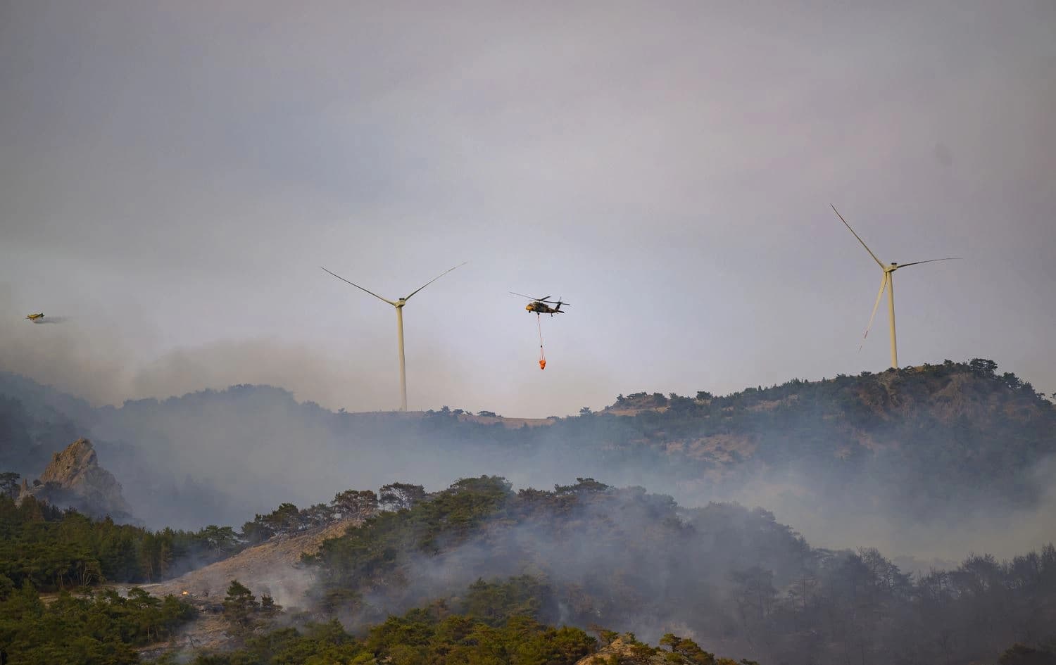 A helicopter carrying water to fight a forest fire with windmills in the background in Turkey's western province of Izmir on August 17, 2024. Photo: Yasin Akgul/AFP