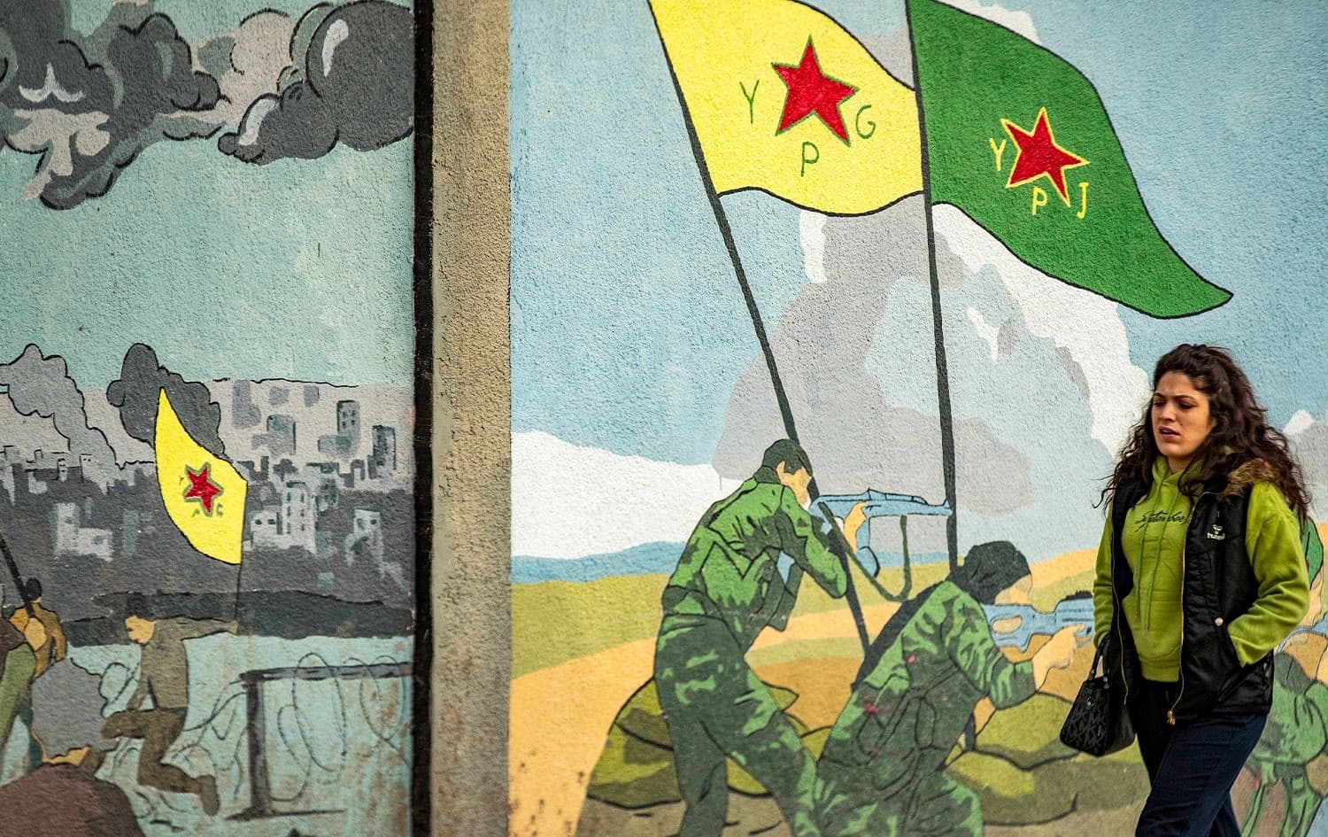 A woman walks past a mural on a wall depicting fighters of the Kurdish People's Protection Units (YPG) and Women's Protection Units (YPJ) in combat along the front lines in Syria's northeastern city of Qamishli on December 16, 2024. Photo: Delil Souleiman/AFP