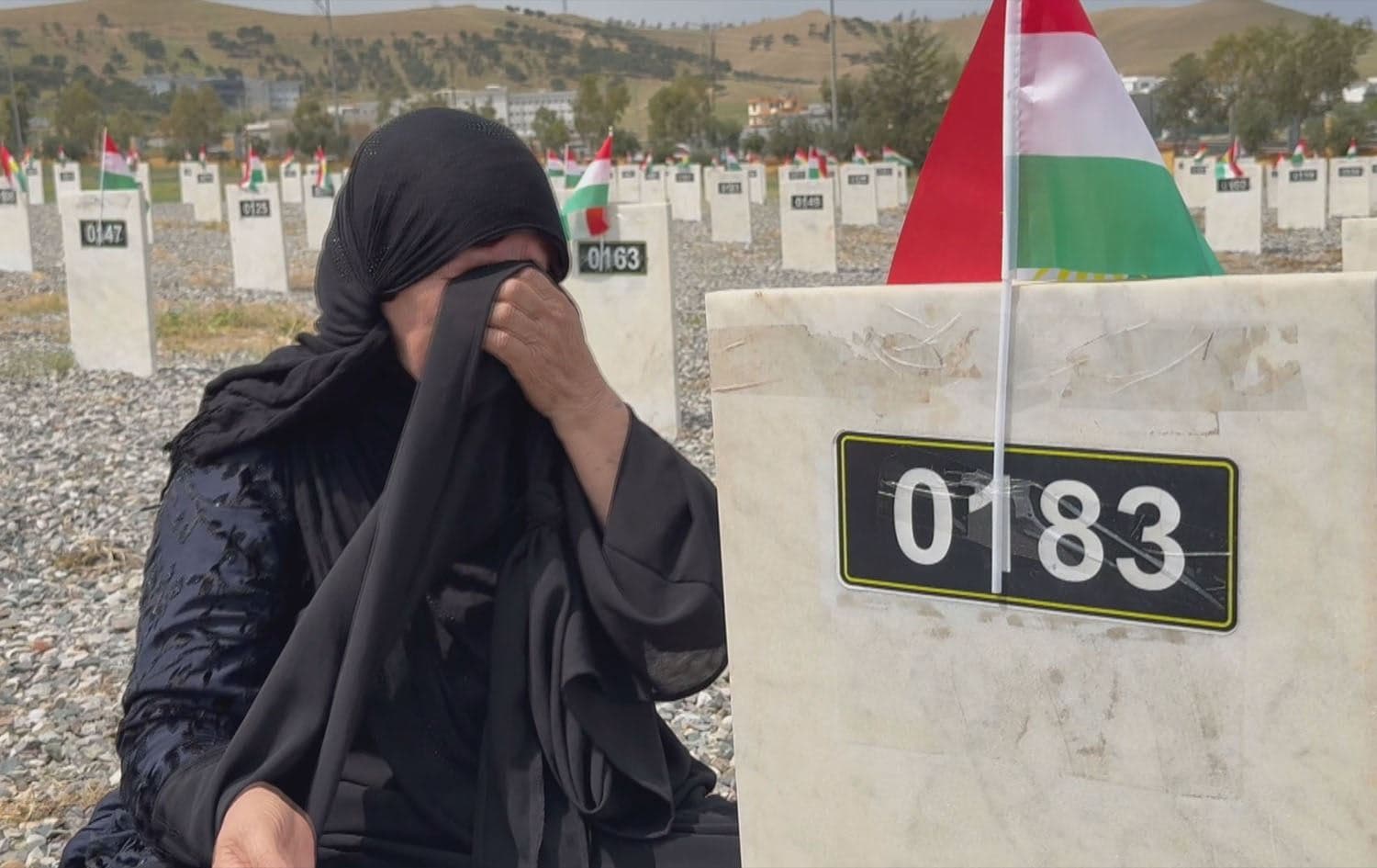 An unnamed gravestone of the remains of an Anfal victim marked ‘0103’ with the crying family of an Anfal victim at the Anfal Memorial Monument and graveyard in Sulaimani’s Chamchamal district on April 15, 2025. Photo: Rudaw