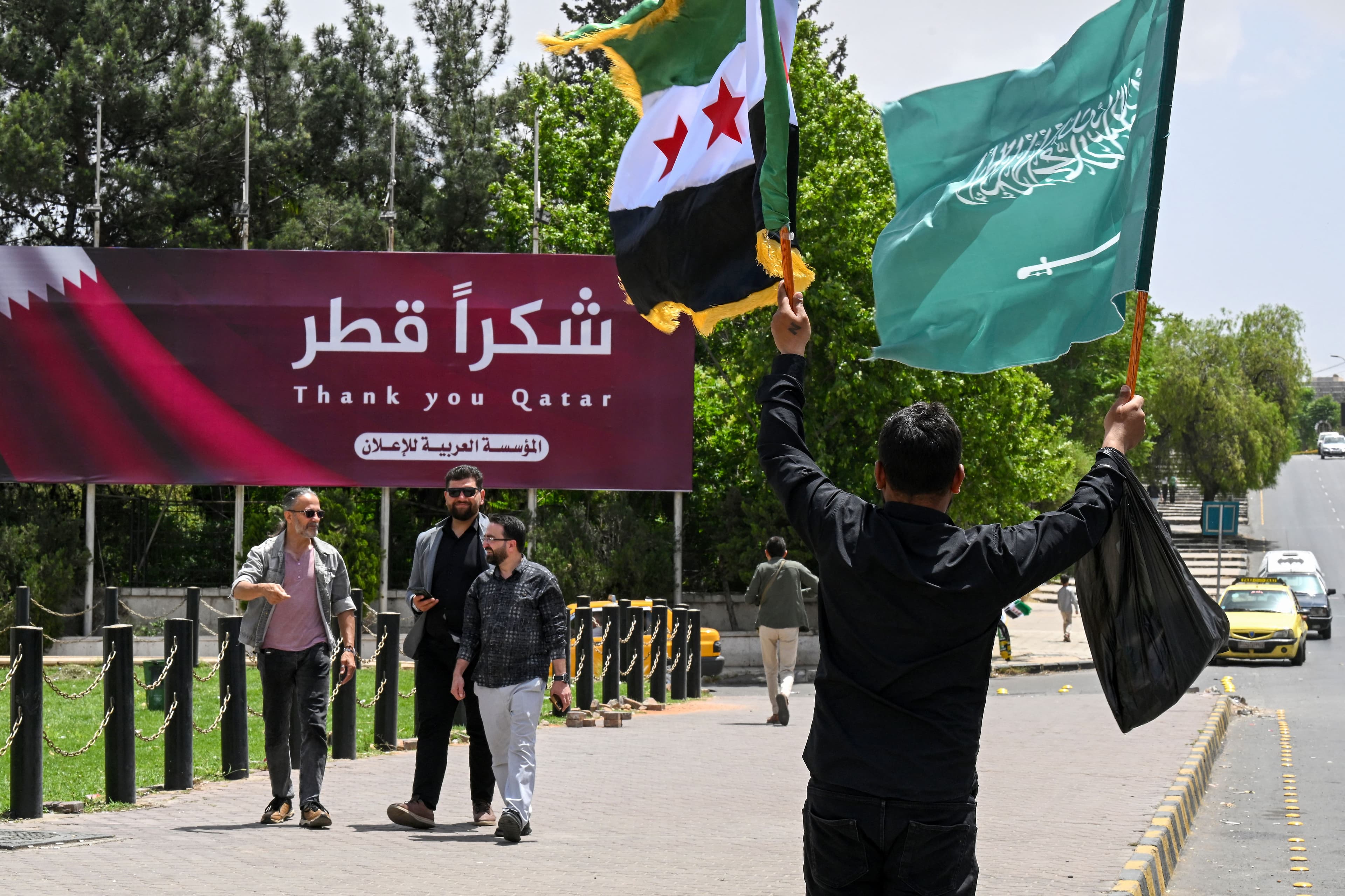 A man carries the Syrian and the Saudi flags past a billboard thanking Qatar, in Damascus on May 14, 2025. Photo: AFP