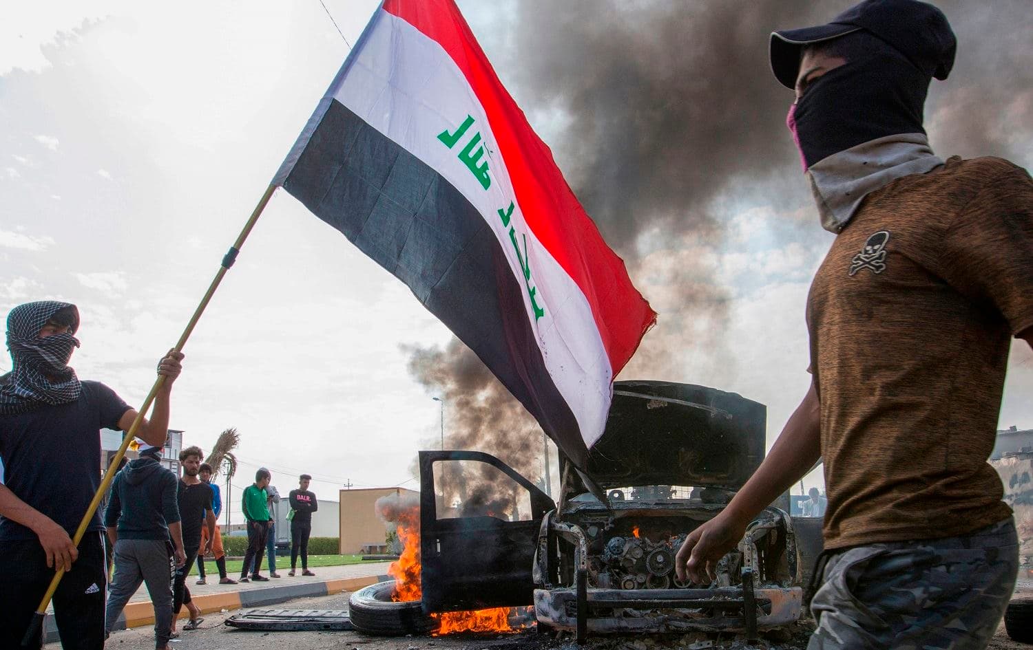 Iraqi demonstrators stand next to the smoking remains of an Iraqi anti-riot vehicle during a demonstration in the southern city of Basra, November 24, 2019. Photo: Hussein Faleh / AFP