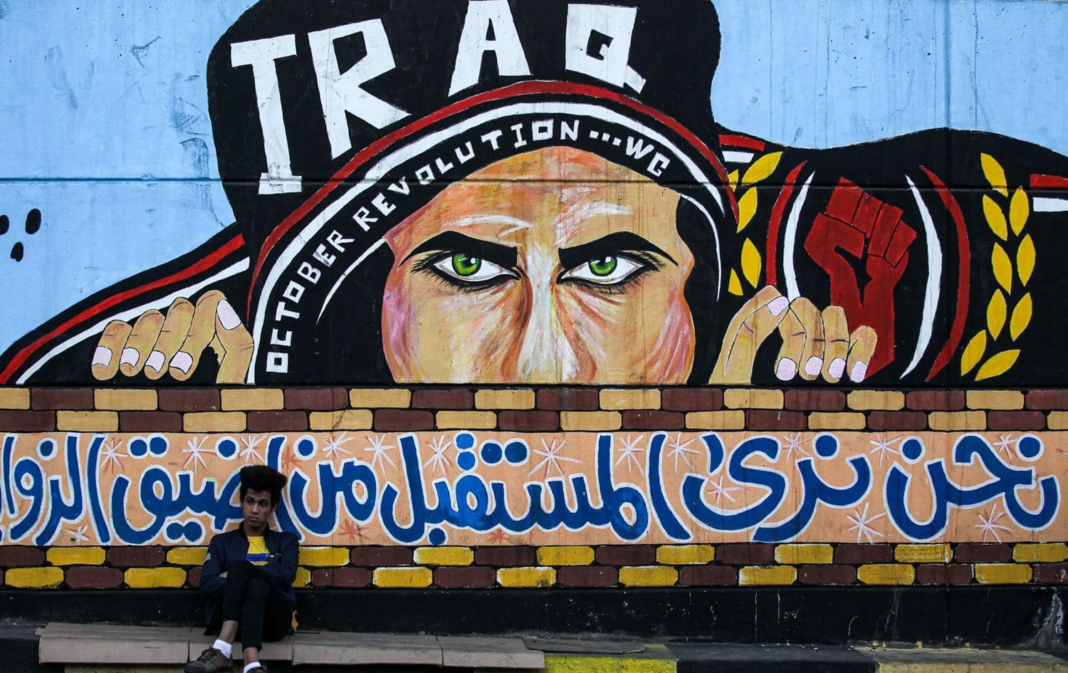 An Iraqi youth sits leaning on a wall with a graffiti mural showing a protester with text in Arabic reaing "we see the future from the narrowest angles", at the anti-government sit-in in the capital's Tahrir Square on December 24, 2019. Photo: Ahmad al-Rubaye