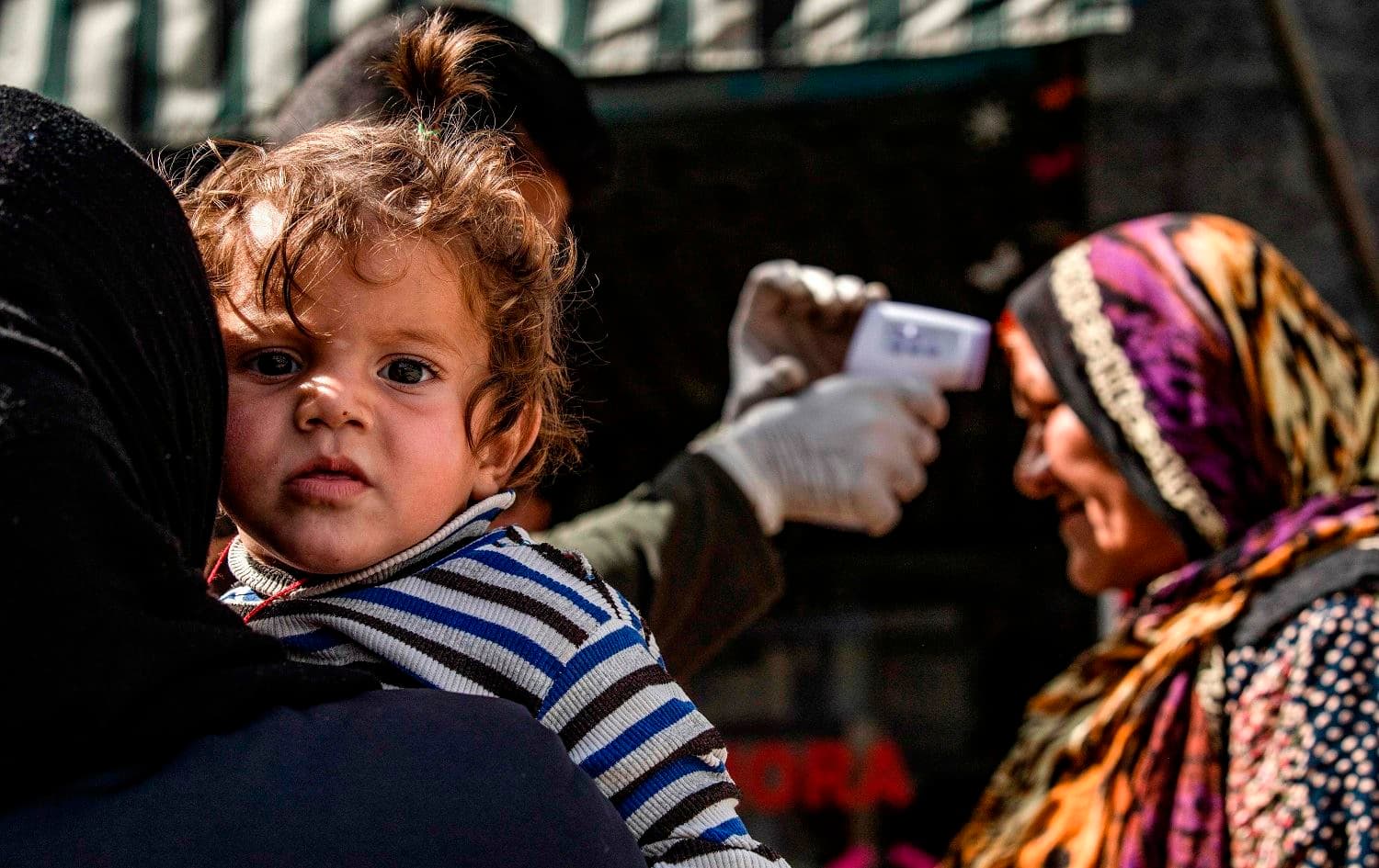 A child looks on as a member of the Asayesh checks a woman's temperature in the Kurdish-majority city of Qamishli in Syria's northeastern Hasakah province, on March 23, 2020.Photo:Delil Souleiman/AFP
