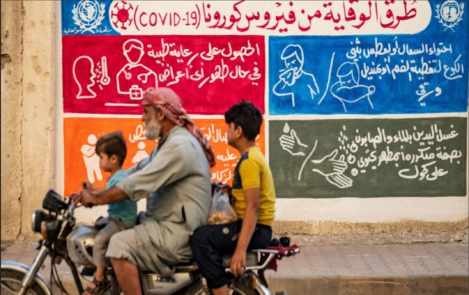 Syrians in the northeast city of Qamishli ride a motorbike past a mural with instructions on protection from COVID-19 in August 2020. Photo: AFP
