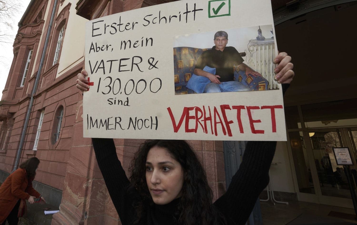 Serda Alshehabi holds a banner in front of the Higher Regional Court in Koblenz, Germany, during an earlier part of the trial on February 24, 2021. The translation reads, "First step. But, my father and 130,000 are still under arrest." Photo: Thomas Frey/AP