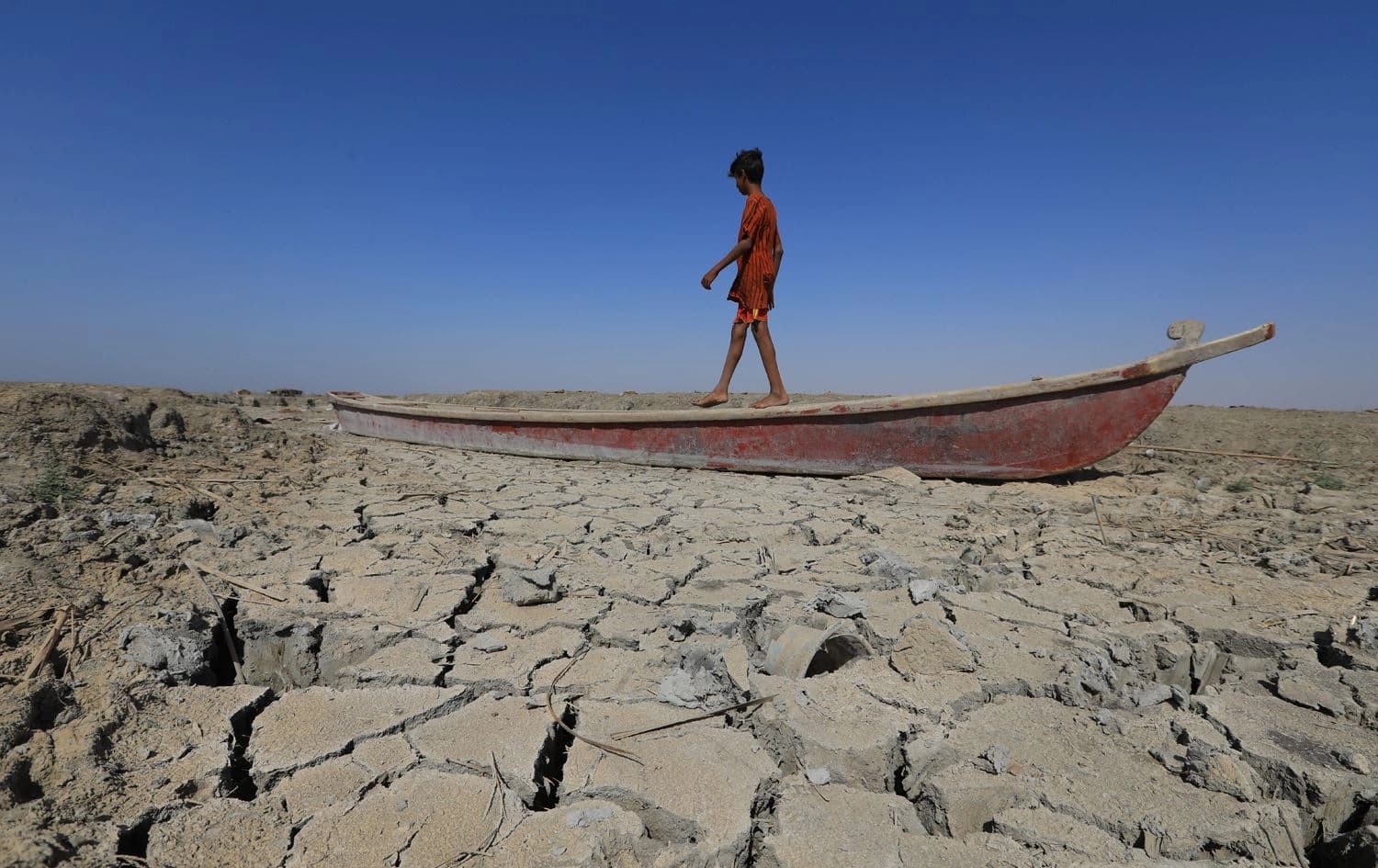 A boy walks on a boat left lying on the dried-up bed of a section of Iraq's receding southern marshes of Chibayish in Dhi Qar province on June 28, 2022. Photo: Asaad Niazi/AFP