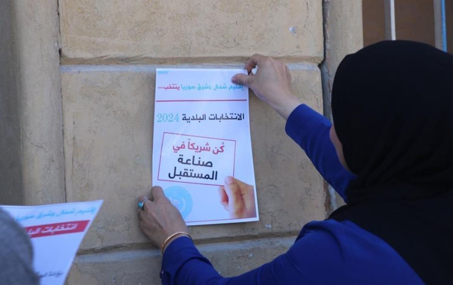A woman hangs a poster on a wall in Rojava, encouraging people to take part in elections. Photo: AA/file