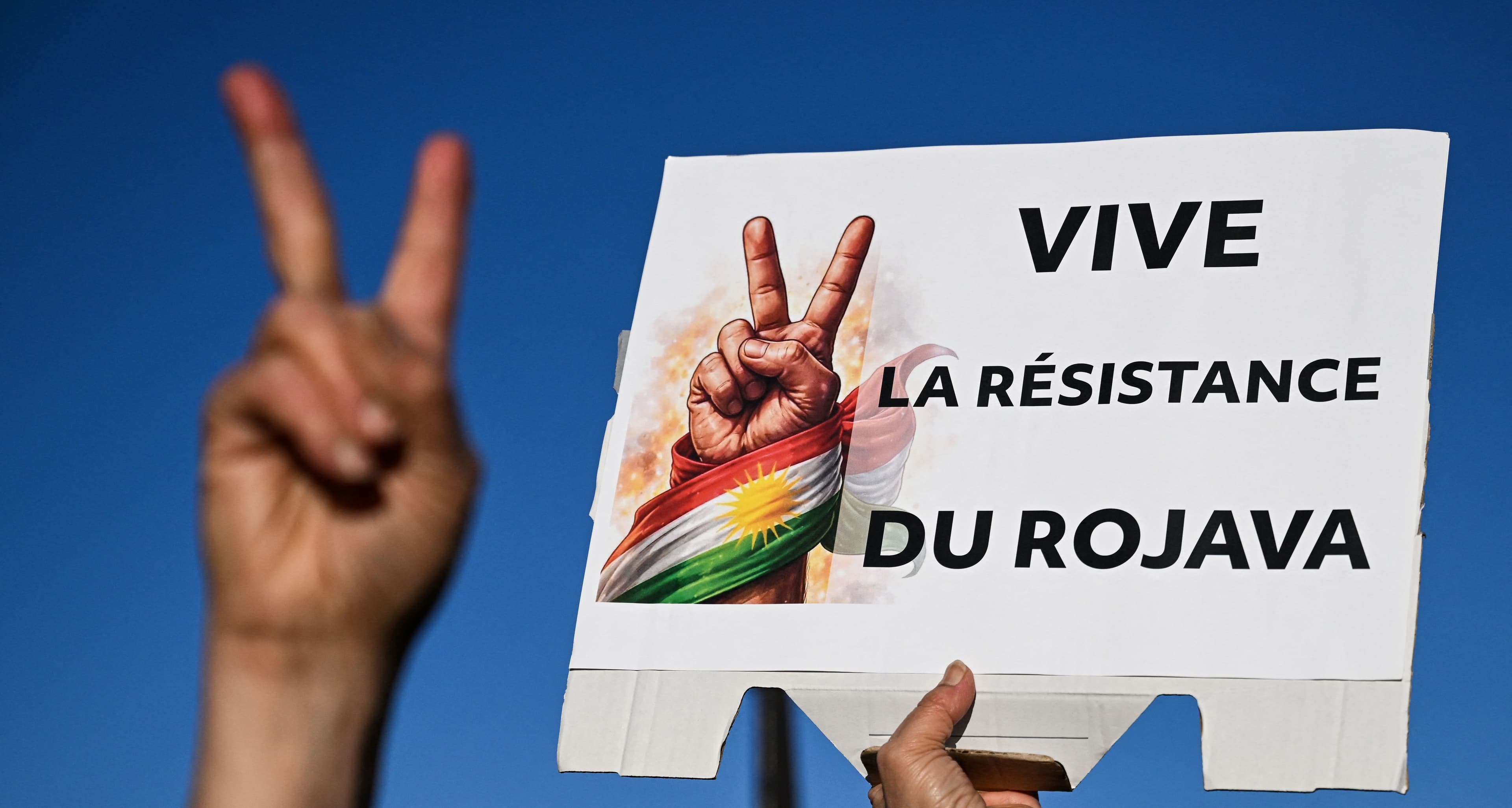 A protester flashes a peace sign with his fingers in front of a placard reading "Long live the resistance of Rojava", which is the Kurdish name for Kurdish-majority regions of Syria, during a demonstration in solidarity with Kurds in northern Syria, in central Paris, on January 24, 2026. Photo: AFP